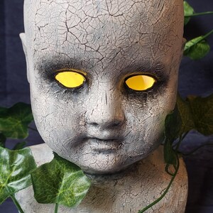 May include: A white ceramic doll's head with cracked paint and glowing yellow eyes. The doll's head is on a grey base with green vines.