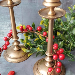 May include: Two gold metal candle holders with a decorative design. The candle holders are on a gray surface with red berries and green leaves.
