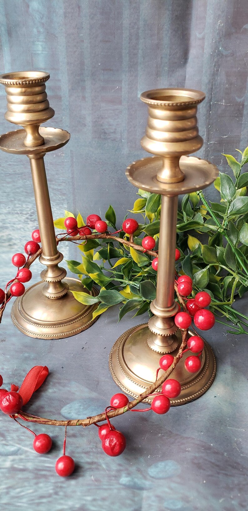 May include: Two gold metal candle holders with a decorative design. The candle holders are on a gray surface with red berries and green leaves.