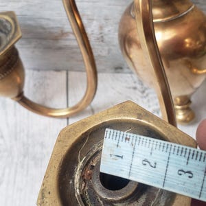 May include: Antique brass candle holder with a curved arm and hexagonal base. The base measures approximately 1 inch across. The candle holder has a decorative, vintage design and is set against a light wood background.