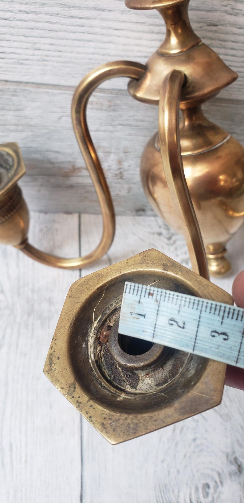 May include: Antique brass candle holder with a curved arm and hexagonal base. The base measures approximately 1 inch across. The candle holder has a decorative, vintage design and is set against a light wood background.