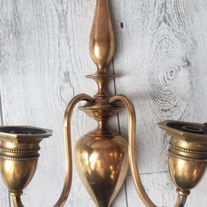 May include: A vintage brass wall sconce with two candle holders. The sconce has a central, elongated body with curved arms extending to the candle cups. The brass has a warm, aged patina, adding to its antique charm.