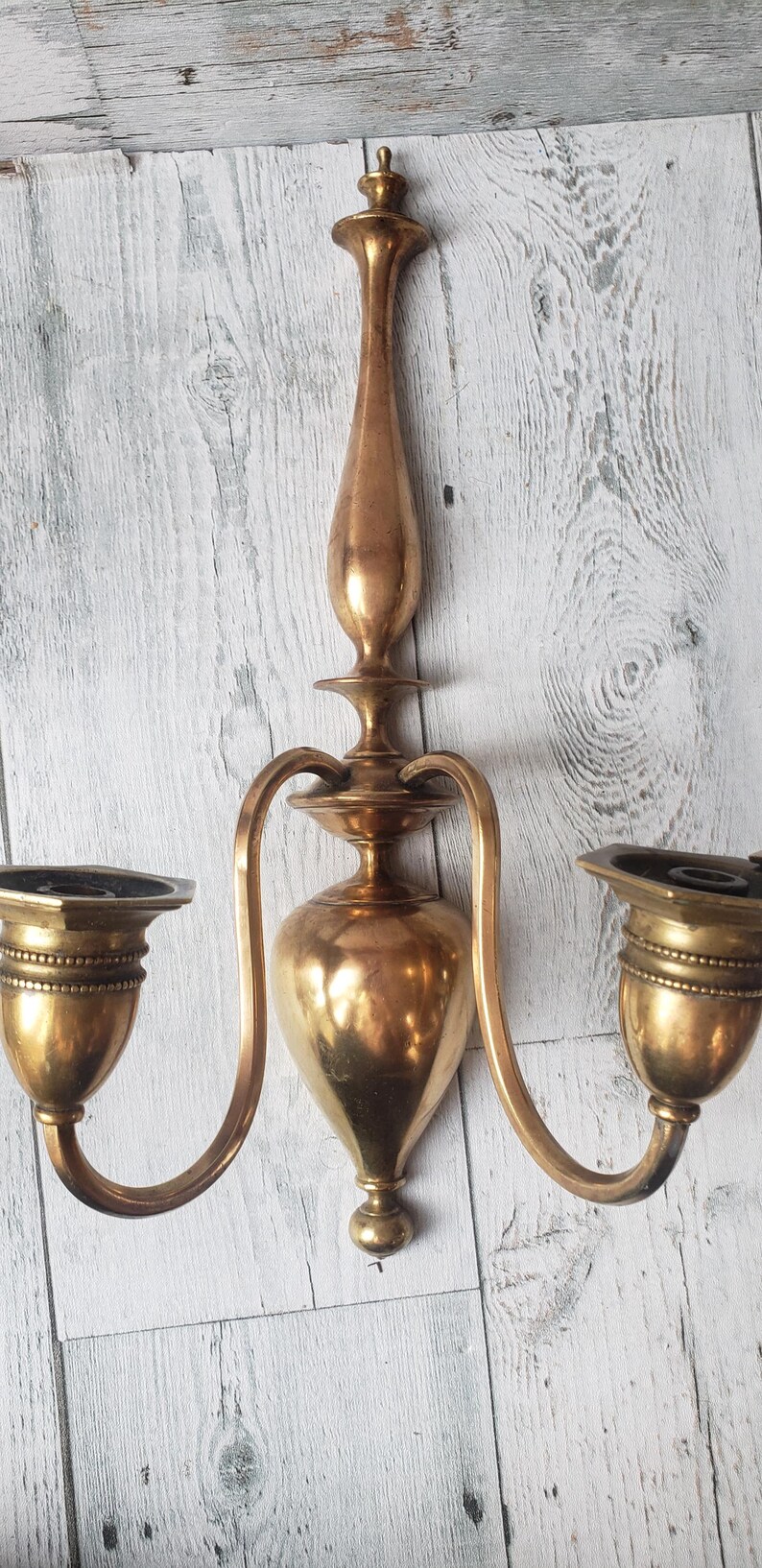May include: A vintage brass wall sconce with two candle holders. The sconce has a central, elongated body with curved arms extending to the candle cups. The brass has a warm, aged patina, adding to its antique charm.