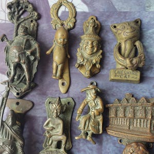 May include: A collection of nine antique brass door knockers featuring various figures including a standing man, a small child, a jester, a Cheshire cat, a seated figure, a building labelled The Ancient House, and a bust labelled King Charles.