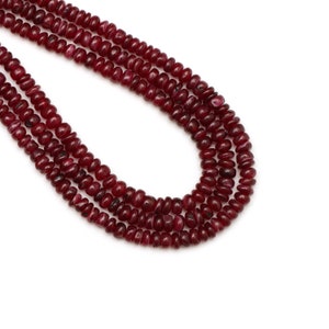 May include: Several strands of deep red gemstone beads, possibly rubies, are shown against a white backdrop. The small, rounded beads have a polished finish. The arrangement suggests jewellery making or crafting.