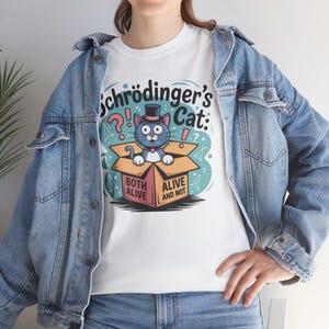 Könnte beinhalten: Weißes T-Shirt mit einer Cartoon-Illustration von Schrödingers Katze. Das Design zeigt eine blaue Katze in einer Kiste mit Zylinder, mit dem Text "Schrödinger's Cat" und den Sätzen "Both Alive" und "Alive and Not".