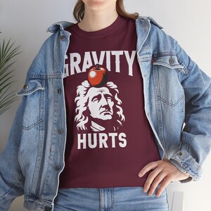 Könnte beinhalten: Ein weinrotes T-Shirt mit einer Grafik, die einen roten Apfel über einer weißen Illustration eines Männerkopfes zeigt, mit den weißen Worten "GRAVITY" und "HURTS". Die Person trägt eine blaue Jeansjacke.