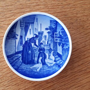 May include: Blue and white ceramic plate featuring a scene of people walking down a street with houses and geese. The plate is decorated with a blue and white floral pattern.