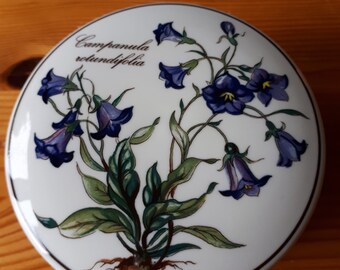 May include: A white ceramic round trinket box with a floral design. The box features a blue and purple flower with green leaves and the text "Campanula rotundifolia".