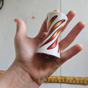 May include: A white hourglass-shaped object with a spiral cutout design revealing a metallic red and copper interior. The object is positioned above a yellow measuring tape that shows a length of approximately 3 inches in total width.