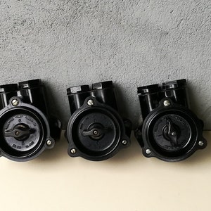 Peut inclure: Trois composants électriques noirs vintage avec des faces circulaires et des ports doubles supérieurs. Chaque composant a un cadran central et est fixé avec des vis. Les composants sont disposés en rangée sur un fond gris texturé.