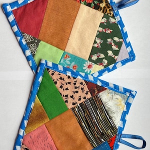Puede incluir: Dos salvamanteles patchwork hechos a mano. Cada salvamanteles presenta una variedad de patrones y colores de tela, incluyendo estampados rojos, verdes, naranjas y florales. Los bordes están ribeteados con un ribete a rayas azules y blancas, y se adjunta un lazo azul para colgar.