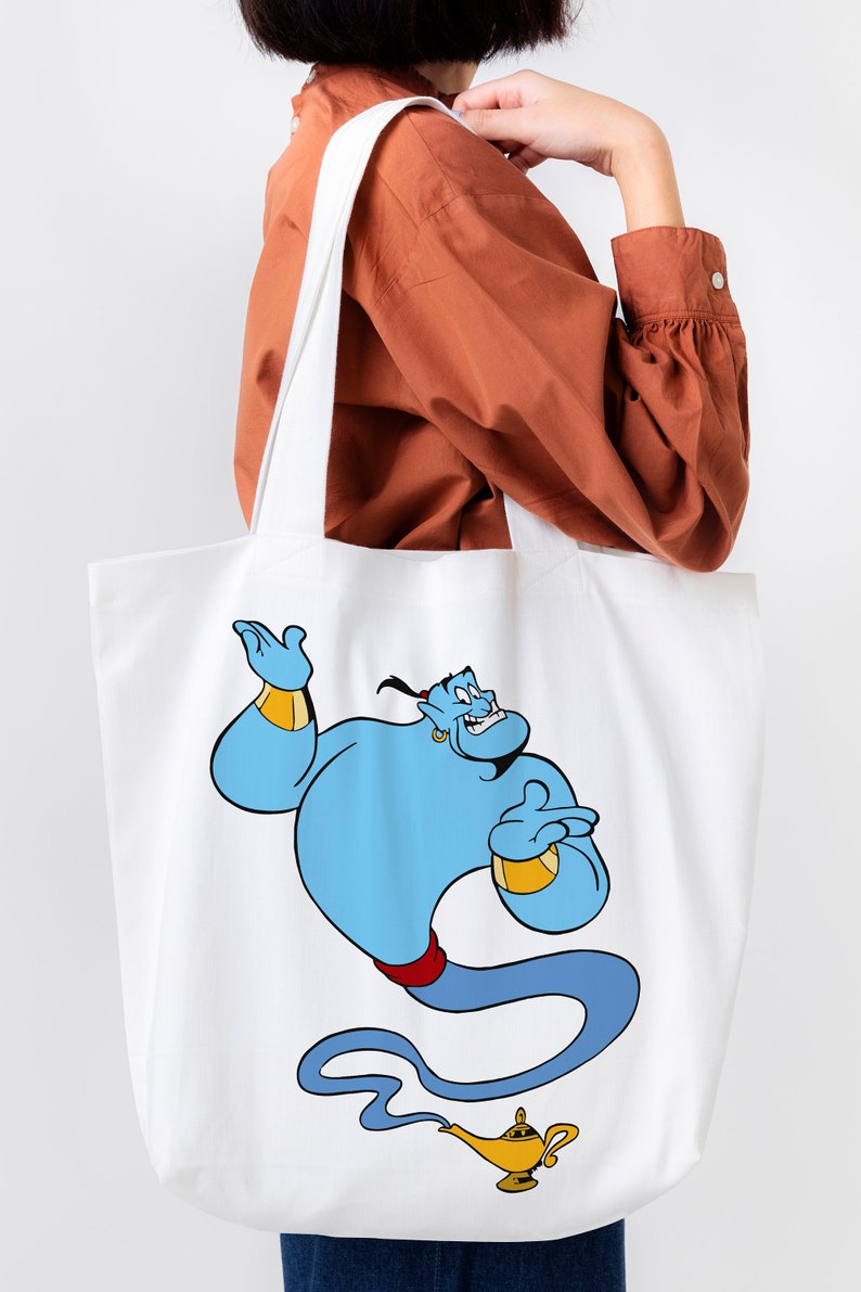 May include: White canvas tote bag with a blue genie character from Disney's Aladdin. The genie is holding a gold lamp and has a mischievous expression.
