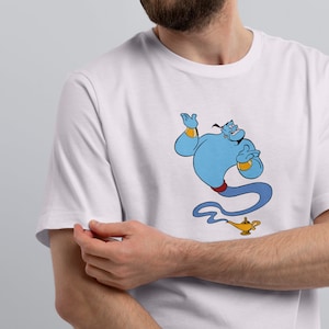 May include: A white t-shirt with a blue genie graphic and a gold lamp. The genie is holding a hand up and smiling. The lamp is on the ground below the genie.