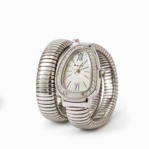 May include: Silver-toned wristwatch with a unique, flexible band. The watch face is oval, with a silver radial pattern and Roman numeral hour markers. The bezel is encrusted with small, clear crystals. The band wraps around the wrist in a coiled design.