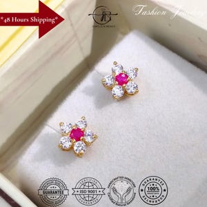 Puede incluir: Un par de pendientes de botón dorados con forma de flor. Cada pendiente presenta una gema rosa central rodeada de gemas blancas, creando un diseño floral. Los pendientes se muestran sobre una superficie blanca dentro de una caja. La imagen también incluye texto.