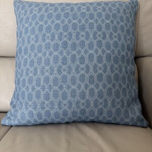 May include: A square throw pillow featuring a medium blue fabric base with a repeating white geometric pattern. The design consists of interconnected rounded arch shapes and circles, creating a consistent, symmetrical grid across the surface.