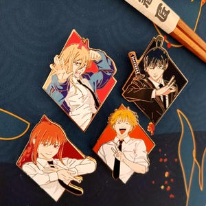 Może przedstawiać: Cztery emaliowane przypinki z postaciami z anime w kształcie rombu z czerwonymi i białymi obramowaniami. Postacie mają różne kolory włosów i wyrazy twarzy. Pałeczki z japońskim tekstem w tle.