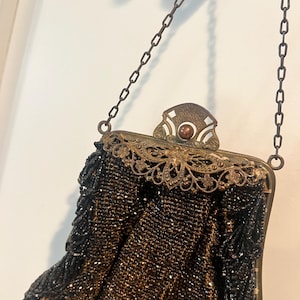 May include: An antique black beaded handbag with a decorative bronze-colored frame and chain handle. The frame features intricate filigree designs and a central embellishment. The bag's beaded body has a fringe detail along the bottom edge.