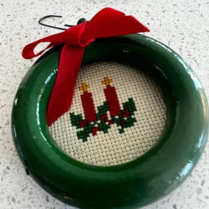 May include: A round, dark green wooden frame containing a cross-stitch embroidery on white fabric. The embroidery depicts two red candles with yellow flames surrounded by green holly leaves. A red ribbon is tied to the top of the frame.