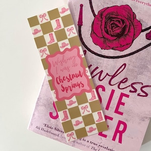 May include: A pink and gold bookmark with a checkered pattern and pink cowboy boots and hats. The bookmark has the text "Wishing I was in Chestnut Springs".