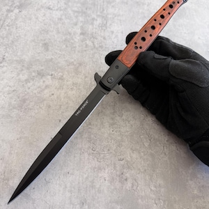 May include: A black stiletto knife with a wooden handle. The handle is a reddish-brown color with several circular cutouts. The blade is black and has the text "TRC-FORCE" printed on it. The knife is open and ready for use.