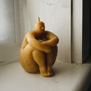 May include: A hand-crafted beeswax candle shaped like a person sitting with their legs crossed. The candle is a light yellow color.