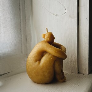 May include: A beige, hand-crafted candle shaped like a person sitting with their arms wrapped around their legs.