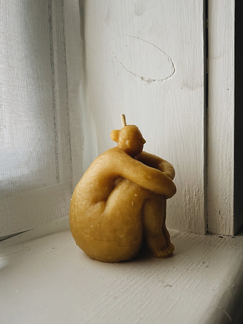 May include: A beige, hand-crafted candle shaped like a person sitting with their arms wrapped around their legs.