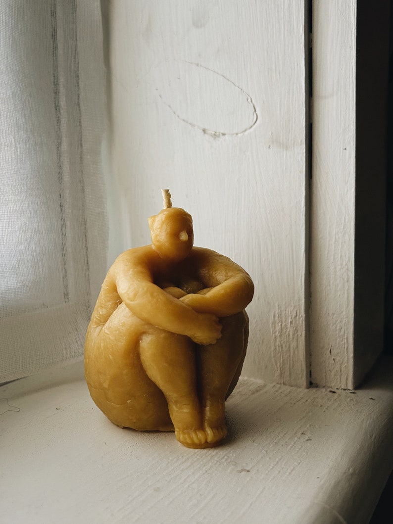 May include: A hand-crafted beeswax candle shaped like a person sitting with their legs crossed. The candle is a light yellow color.