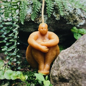 May include: A hand-crafted beeswax candle in the shape of a person sitting with their legs crossed. The candle is a light brown color and has a simple, minimalist design.