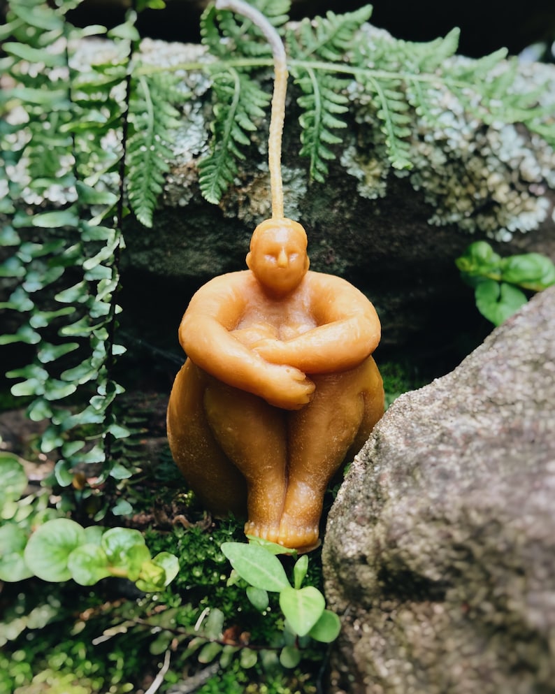 May include: A hand-crafted beeswax candle in the shape of a person sitting with their legs crossed. The candle is a light brown color and has a simple, minimalist design.