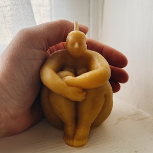 May include: A hand holds a yellow beeswax candle shaped like a person sitting with their legs crossed. The candle has a small wick on top.