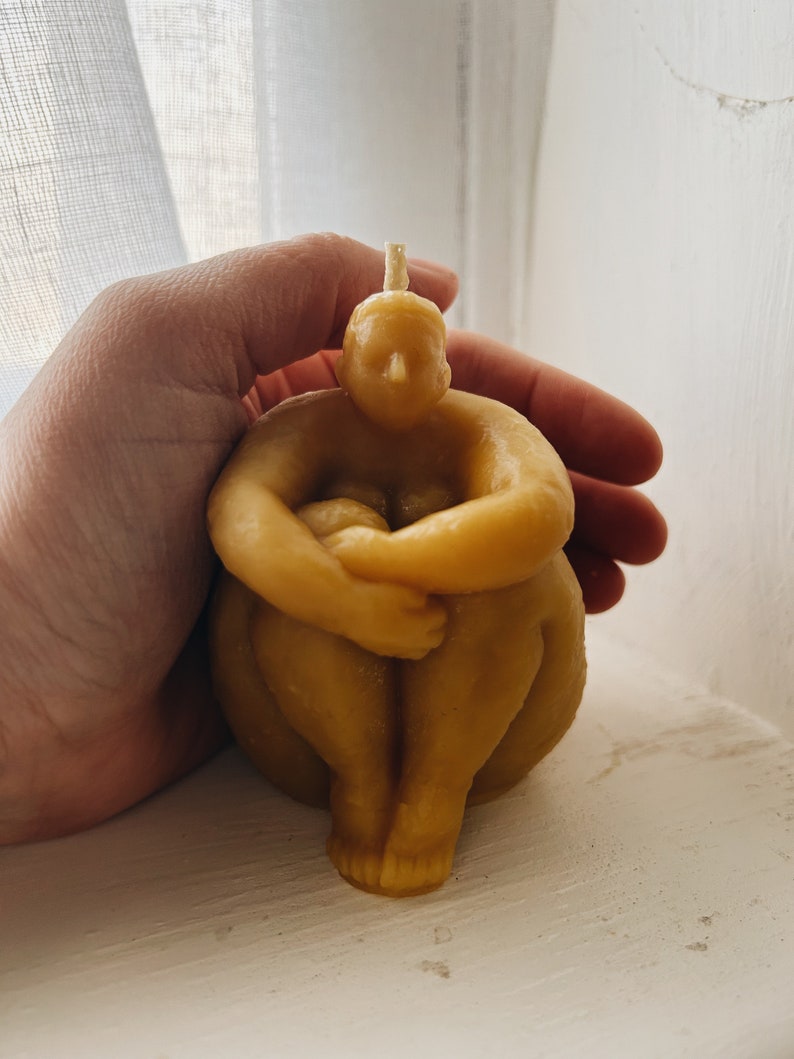 May include: A hand holds a yellow beeswax candle shaped like a person sitting with their legs crossed. The candle has a small wick on top.