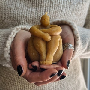 May include: A hand-crafted beeswax candle in the shape of a seated figure. The candle is a light brown color and has a simple, minimalist design.