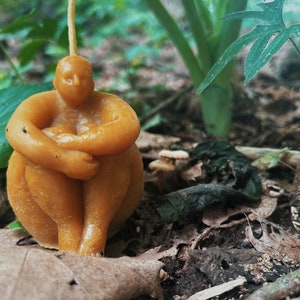 May include: A hand-crafted, honey-colored beeswax candle shaped like a person sitting with their legs crossed. The candle is sitting on a bed of brown leaves and twigs.