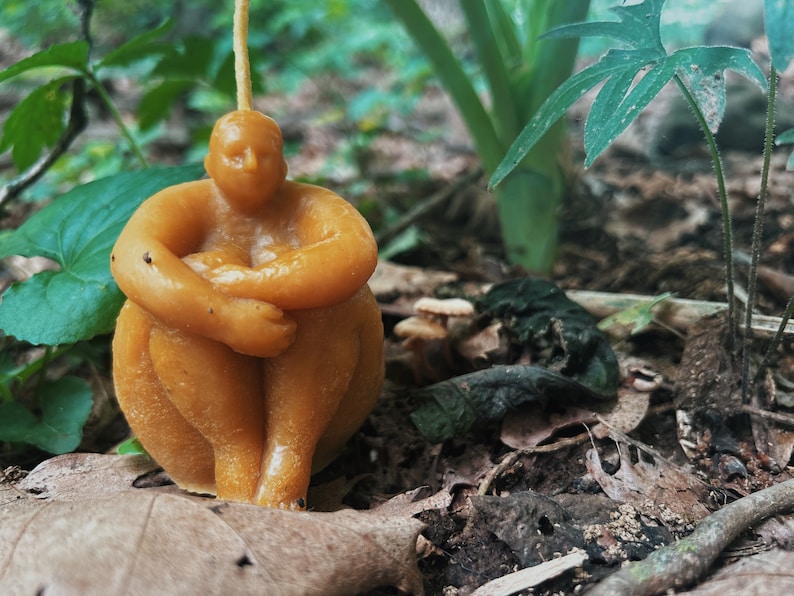 May include: A hand-crafted, honey-colored beeswax candle shaped like a person sitting with their legs crossed. The candle is sitting on a bed of brown leaves and twigs.
