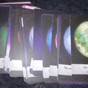 Puede incluir: Un mazo de cartas oráculo rectangulares en abanico, con fondos oscuros, imágenes celestiales, planetas coloridos y bordes blancos en la parte inferior. La carta frontal muestra una luna verde con un rostro y el texto You are good enough.