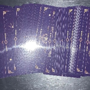 Puede incluir: Una baraja de cartas de tarot rectangulares en abanico, con un fondo morado oscuro y complejos patrones celestiales y geométricos en lámina dorada. La carta de la derecha muestra dos pentagramas y el texto Two of Pentacles.