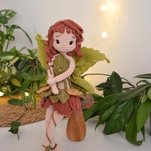 Puede incluir: Una muñeca de hada de ganchillo hecha a mano con cabello rojo, alas verdes y un vestido en forma de hoja. La muñeca está de pie sobre una seta de madera. El hada sostiene una pequeña bolsa verde. Un artículo decorativo caprichoso.