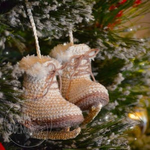 May include: A pair of crocheted ice skate ornaments in cream and brown, with gold-colored blades. The skates are decorated with faux fur trim and tied with brown laces. They hang from a Christmas tree with snowy branches.