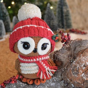 Puede incluir: Una figura de búho de ganchillo que lleva un gorro rojo con un pompón blanco, una bufanda roja y blanca y un gorro rojo con ribete blanco. El búho tiene ojos grandes y redondos y está decorado con una pequeña ramita de acebo. Es una decoración festiva.