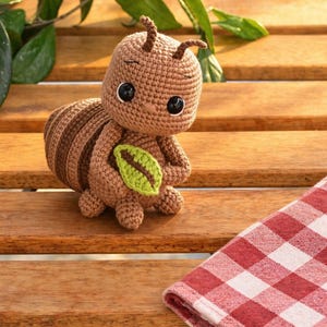 May include: A brown crocheted ant toy with a large head, two antennae, and black plastic eyes. The ant has a segmented body with darker brown stripes and is holding a small, bright green crocheted leaf between its front limbs.