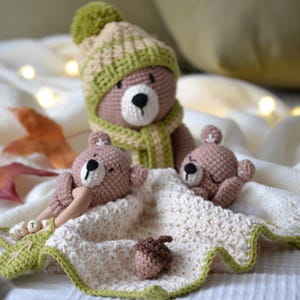 Può includere: Famiglia di orsetti all'uncinetto. L'orso più grande indossa un cappello e una sciarpa verdi e beige. Due orsetti più piccoli riposano su una coperta bianca con bordo verde. Sono presenti anche una ghianda e un massaggiagengive in legno.