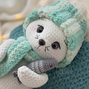 Puede incluir: Un peluche blanco hecho a mano, tejido a ganchillo, con un gorro y bufanda a rayas azul claro y blanco. El juguete de peluche tiene ojos negros, una nariz gris y sostiene un objeto a rayas grises y blancas. Descansa sobre una superficie azul.