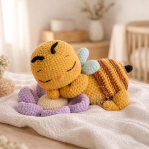 Puede incluir: Un juguete de peluche de abeja de ganchillo en amarillo, marrón y azul claro, descansando sobre una flor morada y crema. La abeja tiene una cara sonriente y está acostada sobre una superficie blanca. El juguete tiene el texto "CAJITA SUENOS".