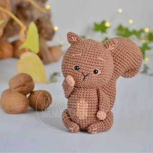May include: A crocheted brown squirrel with a fluffy tail and small, dark eyes. The handmade amigurumi squirrel is posed with one paw near its face. The image also includes walnuts and the text "CAJITA SUEÑOS".