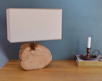 Puede incluir: Una lámpara de mesa de madera con una pantalla rectangular blanca. La base de la lámpara es una gruesa rebanada de madera con veta de madera natural. Un candelero de bronce con una vela blanca está sobre la mesa junto a la lámpara. El libro sobre la mesa dice "Designing a Home & A Life".