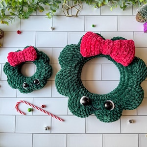 Könnte beinhalten: Zwei grüne gehäkelte Kränze mit roten Schleifen. Die Kränze haben schwarze Knopfaugen und sind mit festlichen Akzenten verziert. Die Kränze sind handgefertigt und eignen sich für die saisonale Dekoration.
