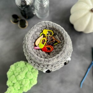 K&ouml;nnte beinhalten: Ein grauer H&auml;kelkorb mit schwarzen Augen und einer wei&szlig;en Nase, gef&uuml;llt mit bunten Garnen und Maschenmarkern. Der Korb steht auf einer grauen Oberfl&auml;che.